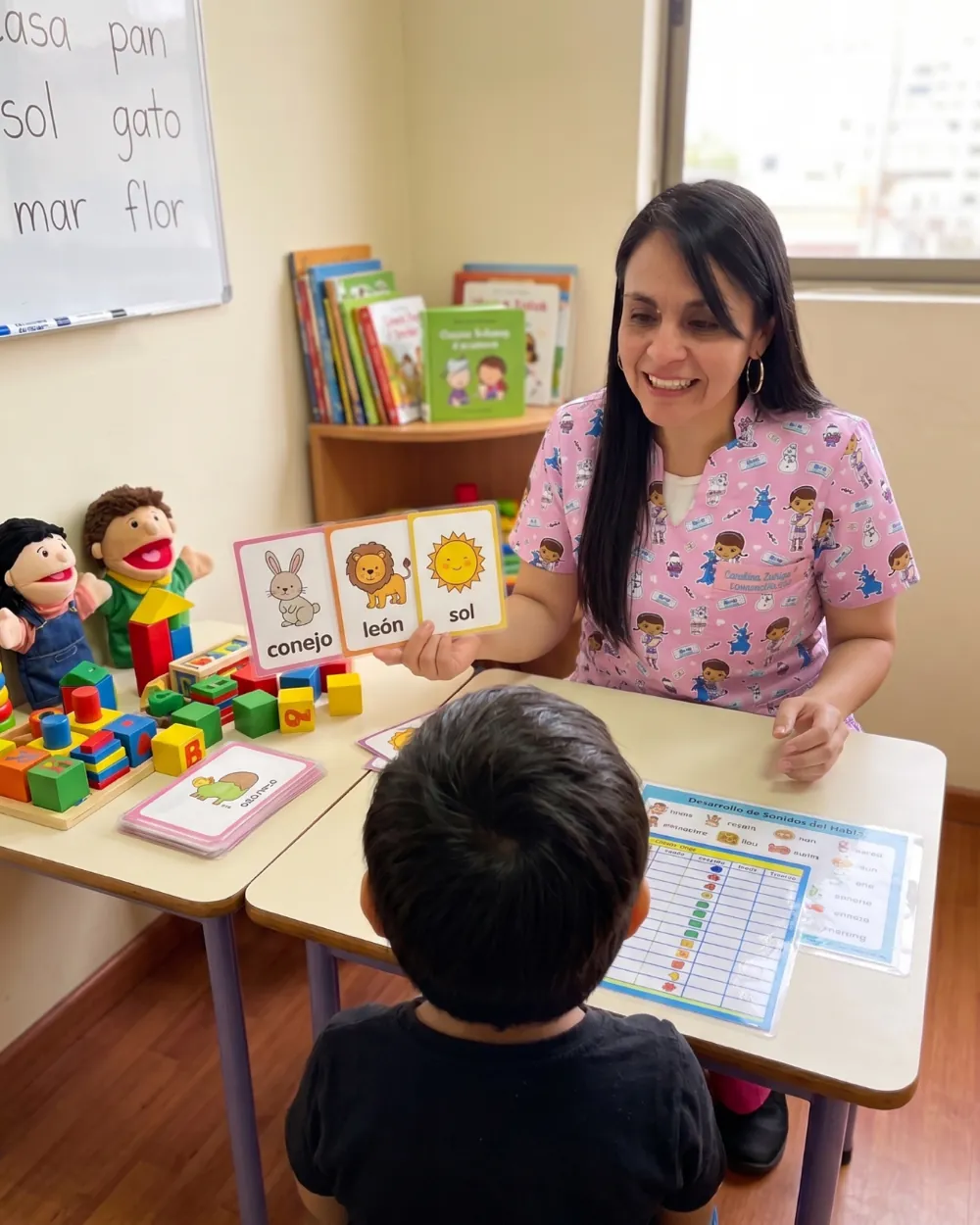 Carolina Zúñiga mostrando tarjetas de vocabulario a un niño, con títeres, bloques y una tabla de desarrollo de los sonidos del habla sobre la mesa