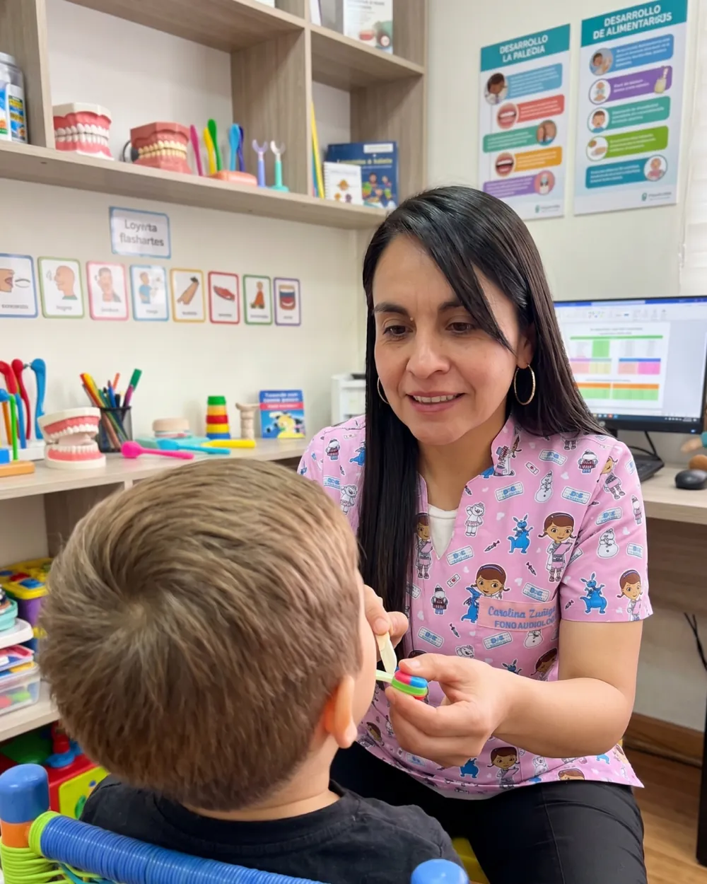 Carolina Zúñiga trabajando la musculatura orofacial de un niño con un depresor, con modelos de arcadas dentales y láminas de desarrollo de la alimentación al fondo